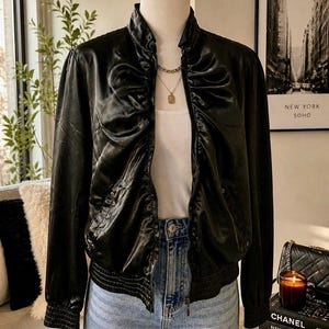 May include: A black satin bomber jacket with a ruched design, a stand-up collar, and ribbed cuffs and hem. The jacket is zipped up and worn over a white tank top and blue jeans. A gold necklace is visible.