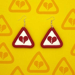 Puede incluir: Un par de pendientes triangulares rojos con fondo blanco y un diseño de corazón roto. Los pendientes están sobre un fondo amarillo.