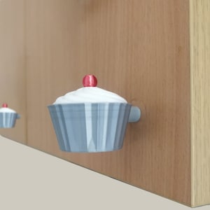 May include: Three grey and white cupcake-shaped cabinet knobs with red centres. The knobs are mounted on a light brown wooden surface.