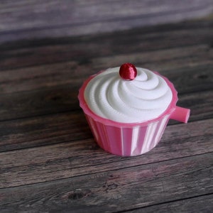 May include: A pink cupcake-shaped container with a white swirl design on top and a small red cherry. The container has a handle and is set on a wooden surface. The cupcake is a decorative item.