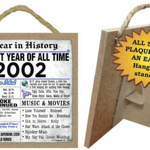 May include: A brown wooden plaque with a newspaper-style design featuring the year 2002 and the text "The Best Year of All Time". The plaque includes a list of events and cultural highlights from the year 2002, including music, movies, and news headlines. The plaque has a hanging rope and a stand.
