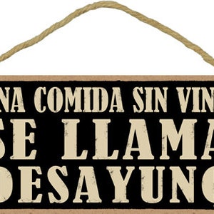 May include: A rustic wooden sign with a black background and white text that reads "UNA COMIDA SIN VINO SE LLAMA DESAYUNO". The sign has a rope hanger.