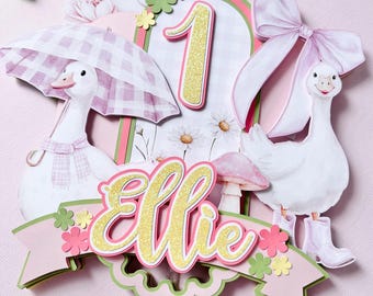 May include: A whimsical cake topper featuring a white goose with a pink gingham umbrella and a bow. The number 1 is displayed with gold glitter, along with the name "Ellie". Pink, green, and white floral accents complete the design.