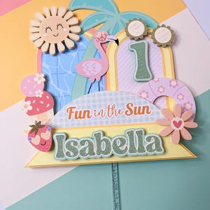 May include: A colorful cake topper with a summer theme. It features a smiling sun, a flamingo, and the number 1. The text "Fun in the Sun" and the name "Isabella" are also included. The topper is attached to a clear stick.