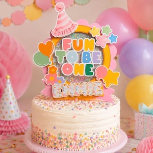May include: A frosted cake with colorful sprinkles on a pink cake stand. The cake is topped with a decorative sign that reads "FUN TO BE ONE" and the name "EMMIE". Balloons and party decorations are in the background.
