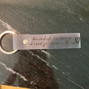 Pode incluir: Um chaveiro de couro cinza com um anel de prata. O chaveiro tem o texto "Drive safe handsome I need you here with me" gravado, juntamente com um pequeno coração.