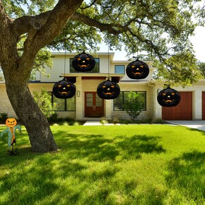 May include: Five black jack-o'-lantern decorations with orange faces hang from tree branches in front of a house. The house has a white exterior with brown trim and a brown door. The jack-o'-lanterns are hanging at different heights and are all facing the camera.