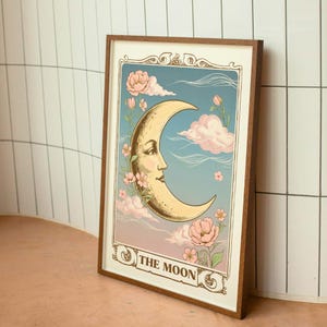 Puede incluir: Una impresión enmarcada de una luna creciente con una cara, rodeada de flores rosas y nubes. El texto "THE MOON" está en la parte inferior de la impresión.