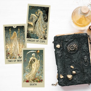 May include: Three tarot cards with illustrations of figures in a fantasy setting, including a knight, a woman, and a skeleton. The cards are labeled "Knight of Cups", "Two of Wan", and "Death". A black, textured book with gold accents is also present.