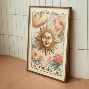 Puede incluir: Una ilustración de carta de tarot de estilo vintage del Sol. La carta presenta un sol dorado con un rostro humano, rodeado de flores rosas y azules. El texto "THE SUN" está impreso en la parte inferior de la carta.