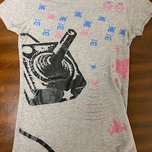 Peut inclure: T-shirt gris avec un motif noir et blanc d'un joystick et d'un vaisseau spatial. Le t-shirt a également des vaisseaux spatiaux et des étoiles pixelisés roses et bleus.