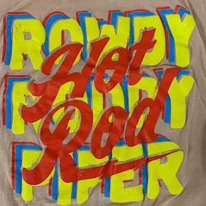 May include: A tan t-shirt featuring the text "ROWDY HOT ROD PIPER". The words are in a layered design, with red text, yellow and blue outlines, creating a 3D effect. The shirt is made of a soft material.