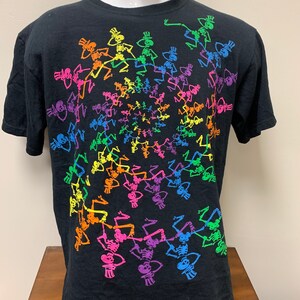 Op de afbeelding: Zwart t-shirt met een spiraalvormig patroon van dansende skeletten in regenboogkleuren. Elk skelet draagt een hoge hoed. Het ontwerp is gecentreerd op de voorkant van het shirt.