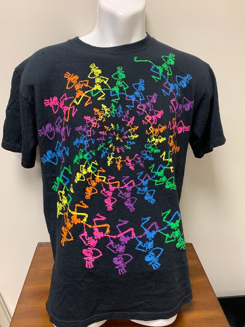 Op de afbeelding: Zwart t-shirt met een spiraalvormig patroon van dansende skeletten in regenboogkleuren. Elk skelet draagt een hoge hoed. Het ontwerp is gecentreerd op de voorkant van het shirt.