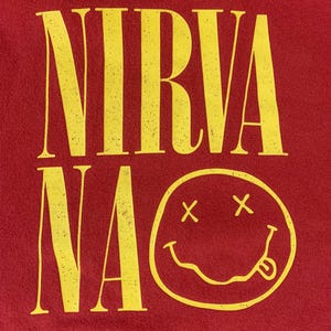 May include: Red hoodie with the word "NIRVANA" in yellow, distressed lettering. Below the word is a yellow smiley face with crossed-out eyes and a tongue sticking out. The hoodie has a hood.