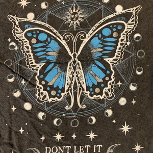 May include: Dark grey t-shirt with a butterfly design in blue and white, set against a celestial backdrop of moons and stars. The text reads "DON'T LET IT PHASE YOU".