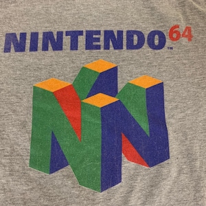 May include: A heather grey t-shirt featuring the blue text NINTENDO 64 with a trademark symbol. Below the text is a large, three-dimensional N64 logo graphic composed of geometric shapes in vibrant shades of blue, green, red, and orange.