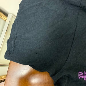 Op de afbeelding: Zwart T-shirt met korte mouwen en een grafisch ontwerp. Het shirt heeft een klein gaatje bij de mouw. Het ontwerp bevat een schedel en andere elementen in blauw en paars.