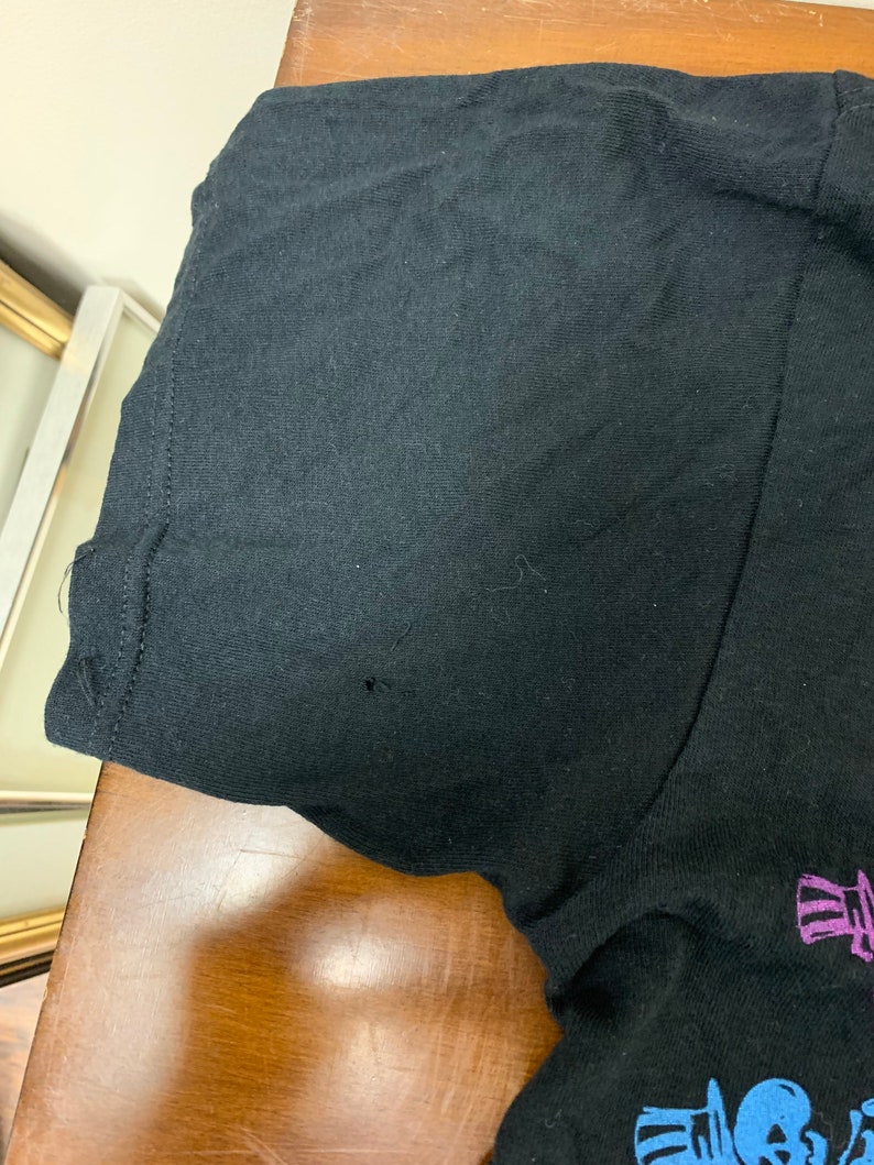 Op de afbeelding: Zwart T-shirt met korte mouwen en een grafisch ontwerp. Het shirt heeft een klein gaatje bij de mouw. Het ontwerp bevat een schedel en andere elementen in blauw en paars.