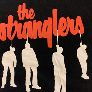 May include: Black t-shirt featuring the band name "The Stranglers" in red, stylized lettering above four white silhouette figures hanging from strings. The figures are in various poses.