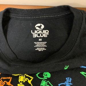 Op de afbeelding: Zwart T-shirt met een kleurrijke skeletafbeelding. Het shirt heeft het "Liquid Blue" logo en maat "M" op de kraag. Gemaakt van 100% voorgekrompen katoen. De afbeelding bevat skeletten in verschillende kleuren.