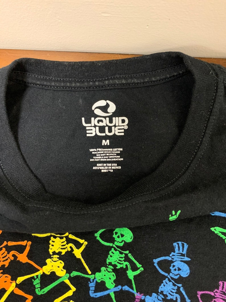 Op de afbeelding: Zwart T-shirt met een kleurrijke skeletafbeelding. Het shirt heeft het "Liquid Blue" logo en maat "M" op de kraag. Gemaakt van 100% voorgekrompen katoen. De afbeelding bevat skeletten in verschillende kleuren.