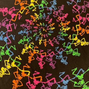 Op de afbeelding: Zwart T-shirt met een spiraalpatroon van dansende skeletten in regenboogkleuren. Elk skelet draagt een hoge hoed. De skeletten zijn in tinten roze, oranje, geel, groen, blauw en paars.