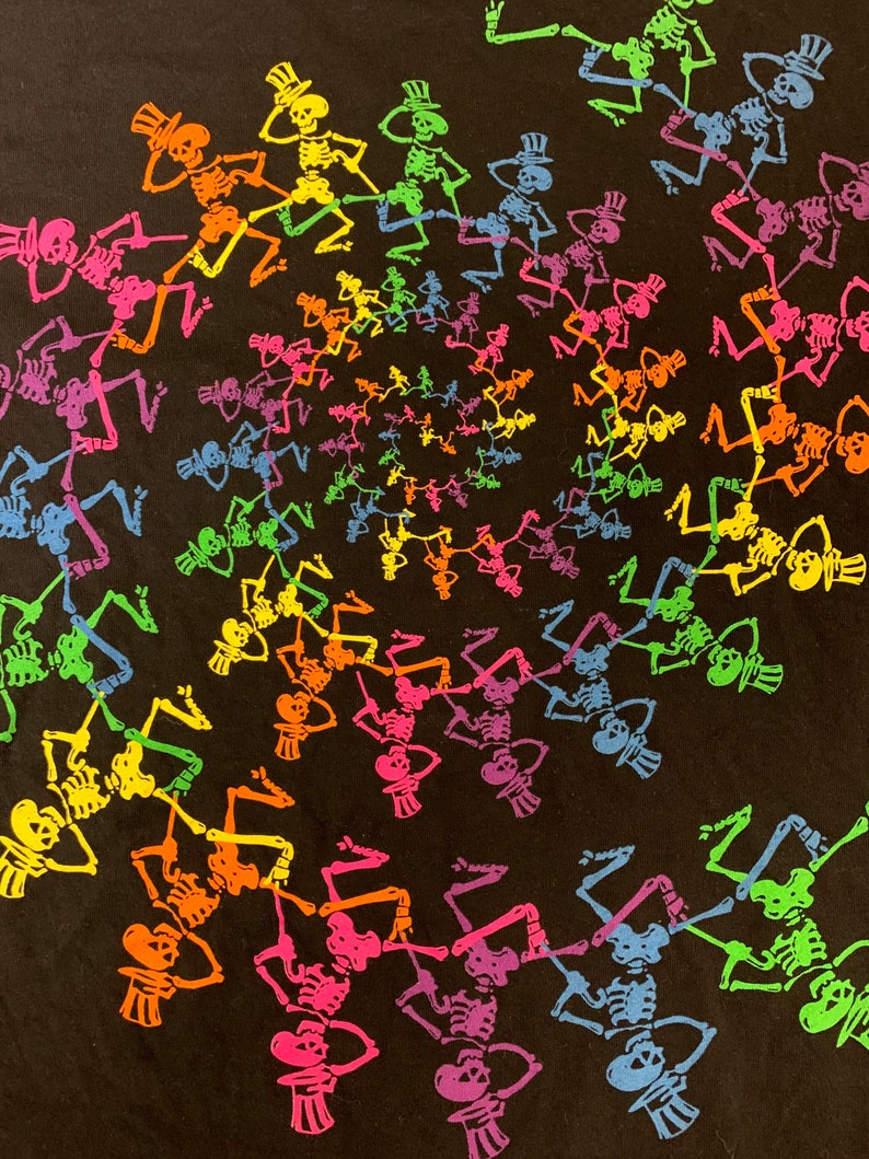 Op de afbeelding: Zwart T-shirt met een spiraalpatroon van dansende skeletten in regenboogkleuren. Elk skelet draagt een hoge hoed. De skeletten zijn in tinten roze, oranje, geel, groen, blauw en paars.