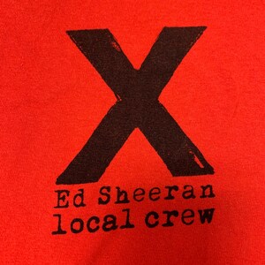 Peut inclure: T-shirt rouge avec un "X" noir et le texte "Ed Sheeran local crew" imprimé sur le devant.