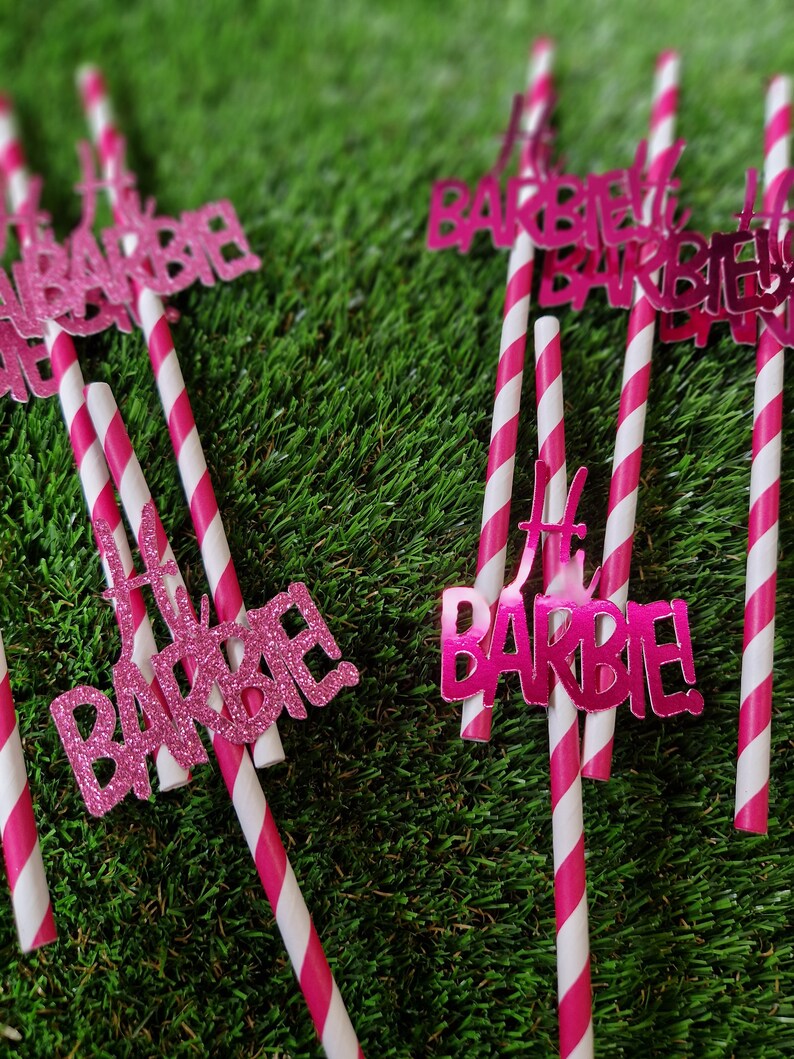 May include: A collection of drinking straws featuring white and pink diagonal stripes. Each straw is topped with a decorative cutout that reads Hi BARBIE! in either a glittery pink finish or a smooth, reflective metallic pink finish.