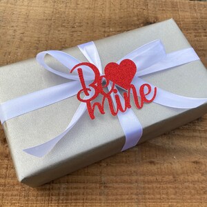 Puede incluir: Una caja de regalo plateada con una cinta de sat&eacute;n blanca y un coraz&oacute;n rojo de purpurina con las palabras "Be Mine" en ella.