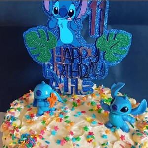 K&ouml;nnte beinhalten: Ein Geburtstagskuchen, verziert mit blauen Stitch-Figuren und einem glitzernden blauen Tortenaufsatz mit der Aufschrift "Happy Birthday" und der Zahl 11. Der Kuchen ist mit wei&szlig;em Zuckerguss und bunten Streuseln bedeckt.
