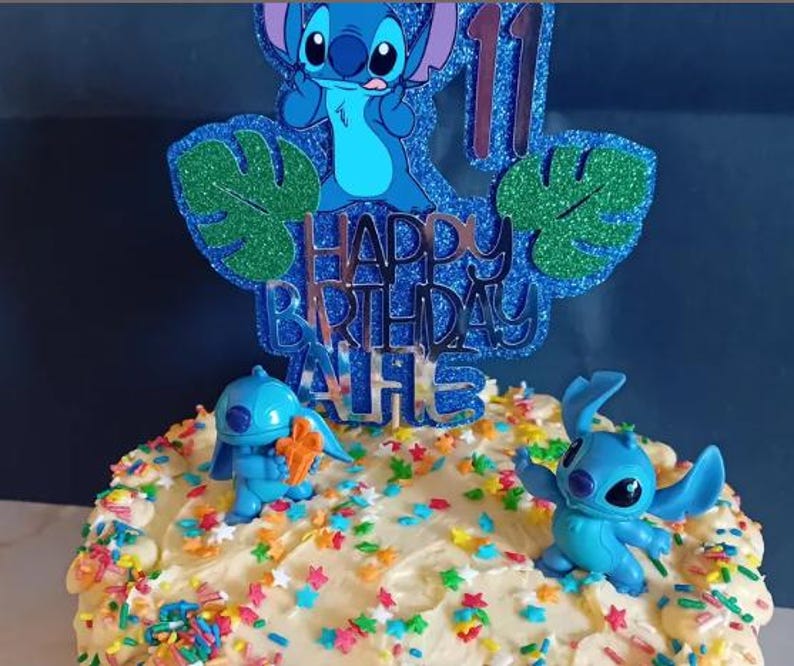 K&ouml;nnte beinhalten: Ein Geburtstagskuchen, verziert mit blauen Stitch-Figuren und einem glitzernden blauen Tortenaufsatz mit der Aufschrift "Happy Birthday" und der Zahl 11. Der Kuchen ist mit wei&szlig;em Zuckerguss und bunten Streuseln bedeckt.