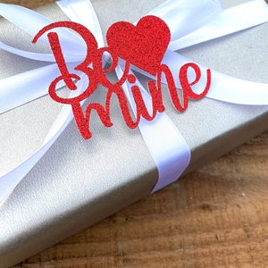 Puede incluir: Una caja de regalo plateada con una cinta de sat&eacute;n blanca y un coraz&oacute;n rojo de purpurina con las palabras "Be mine" en ella.