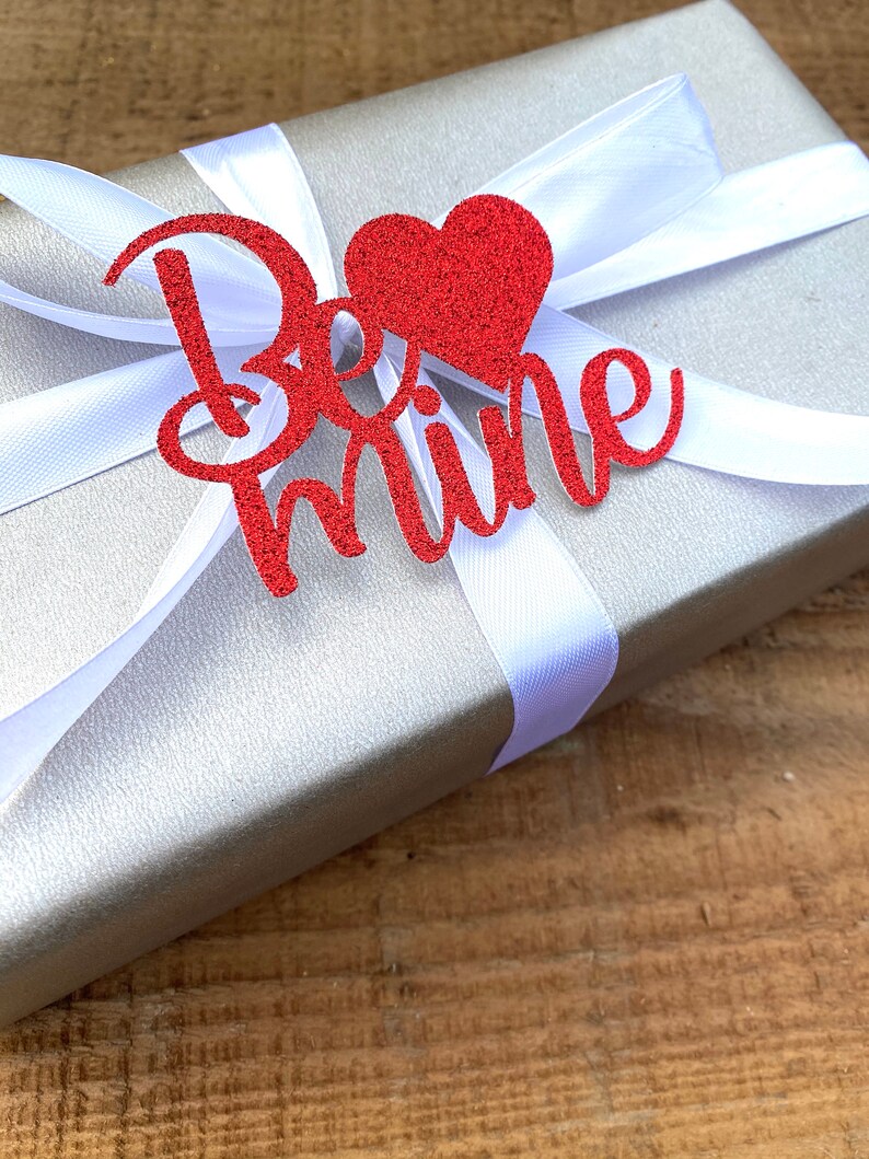 Puede incluir: Una caja de regalo plateada con una cinta de sat&eacute;n blanca y un coraz&oacute;n rojo de purpurina con las palabras "Be mine" en ella.