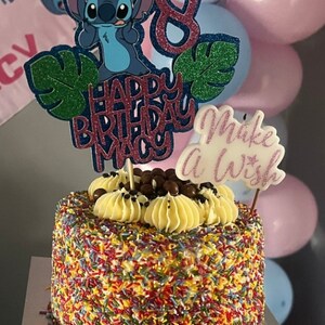 K&ouml;nnte beinhalten: Ein Geburtstagskuchen mit bunten Streuseln, belegt mit gelber Buttercreme, Schokoladenbonbons und einer Stitch-Kuchendekoration. Der Kuchen hat einen Aufsatz mit der Aufschrift "Happy Birthday Macy" und "Make a Wish". Rosa und blaue Luftballons sind im Hintergrund.