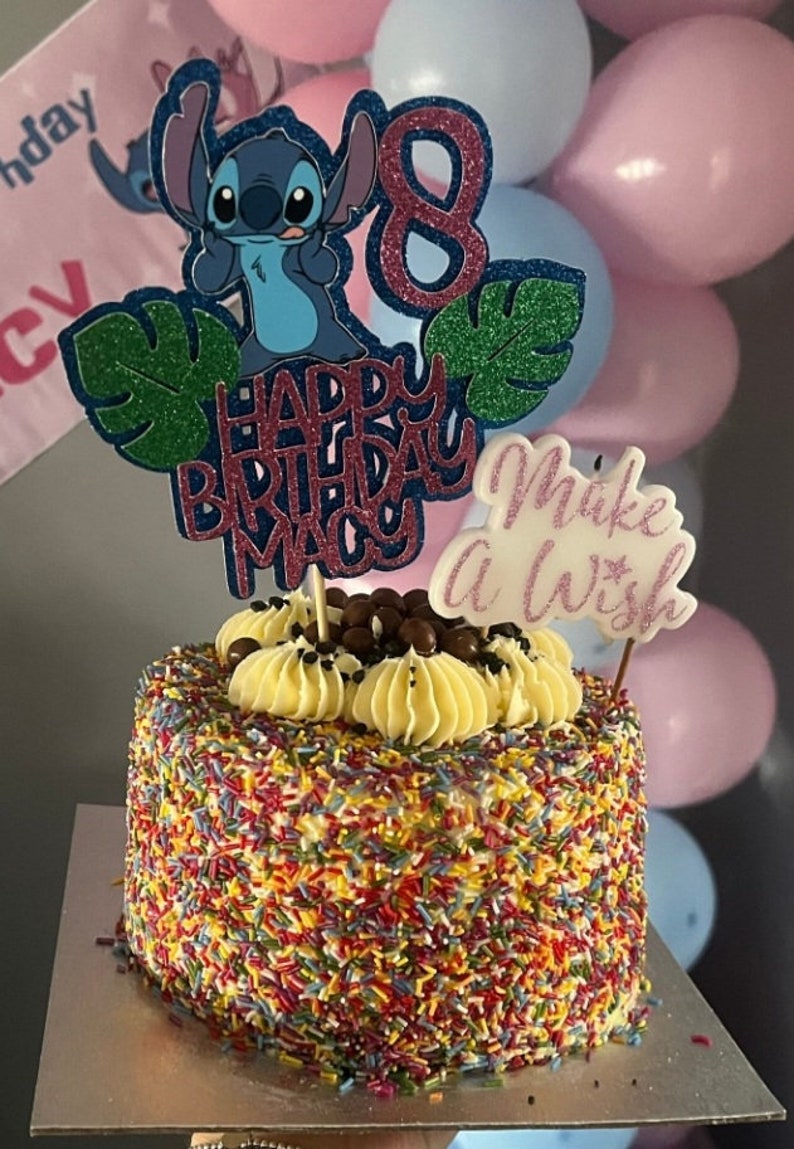 K&ouml;nnte beinhalten: Ein Geburtstagskuchen mit bunten Streuseln, belegt mit gelber Buttercreme, Schokoladenbonbons und einer Stitch-Kuchendekoration. Der Kuchen hat einen Aufsatz mit der Aufschrift "Happy Birthday Macy" und "Make a Wish". Rosa und blaue Luftballons sind im Hintergrund.