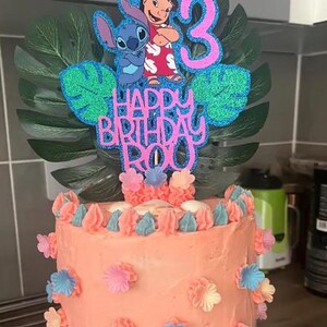K&ouml;nnte beinhalten: Eine Geburtstagstorte mit korallenfarbenem Zuckerguss, verziert mit rosa, blauen und wei&szlig;en Blumenakzenten. Die Torte ist mit einem glitzernden Tortenaufsatz mit den Figuren Stitch und Lilo, der Zahl 3 und dem Schriftzug "Happy Birthday Roy" versehen.