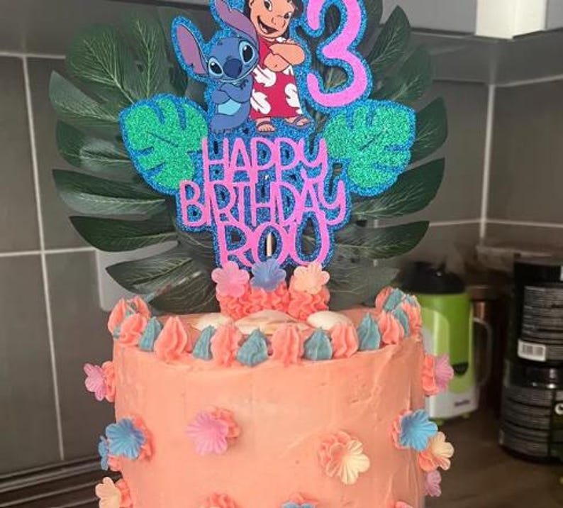 K&ouml;nnte beinhalten: Eine Geburtstagstorte mit korallenfarbenem Zuckerguss, verziert mit rosa, blauen und wei&szlig;en Blumenakzenten. Die Torte ist mit einem glitzernden Tortenaufsatz mit den Figuren Stitch und Lilo, der Zahl 3 und dem Schriftzug "Happy Birthday Roy" versehen.