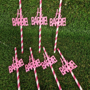 May include: Seven drinking straws with white and pink diagonal stripes, each featuring a pink glittery cutout topper that reads Hi BARBIE! in a stylized script and block font. The straws are arranged in two rows against a green textured surface.