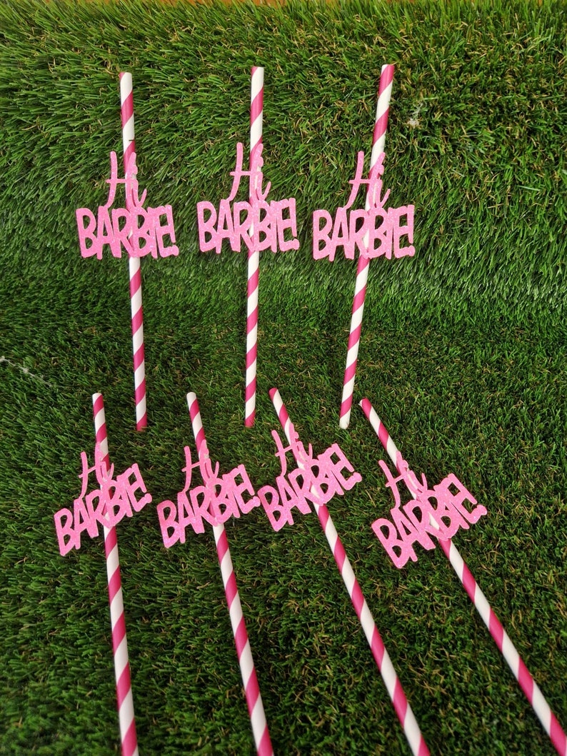 May include: Seven drinking straws with white and pink diagonal stripes, each featuring a pink glittery cutout topper that reads Hi BARBIE! in a stylized script and block font. The straws are arranged in two rows against a green textured surface.