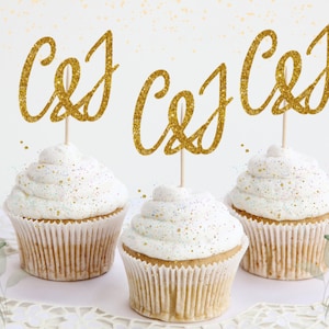 May include: Three cupcakes with white frosting and gold glitter sprinkles topped with gold glitter letter cake toppers that spell out "C&J".