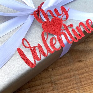 Puede incluir: Una caja de regalo plateada con una cinta de sat&eacute;n blanca y una etiqueta roja con purpurina que dice "Mi Valent&iacute;n".