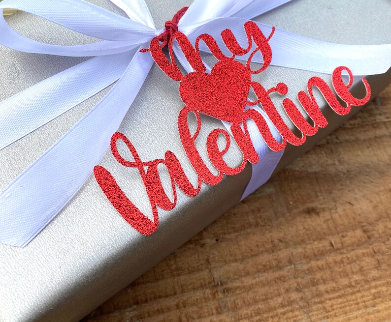 Puede incluir: Una caja de regalo plateada con una cinta de sat&eacute;n blanca y una etiqueta roja con purpurina que dice "Mi Valent&iacute;n".