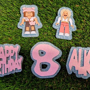 May include: A set of pink and blue glitter cake toppers featuring two cartoon figures, the words "Happy Birthday", the number "8", and the name "Alice".