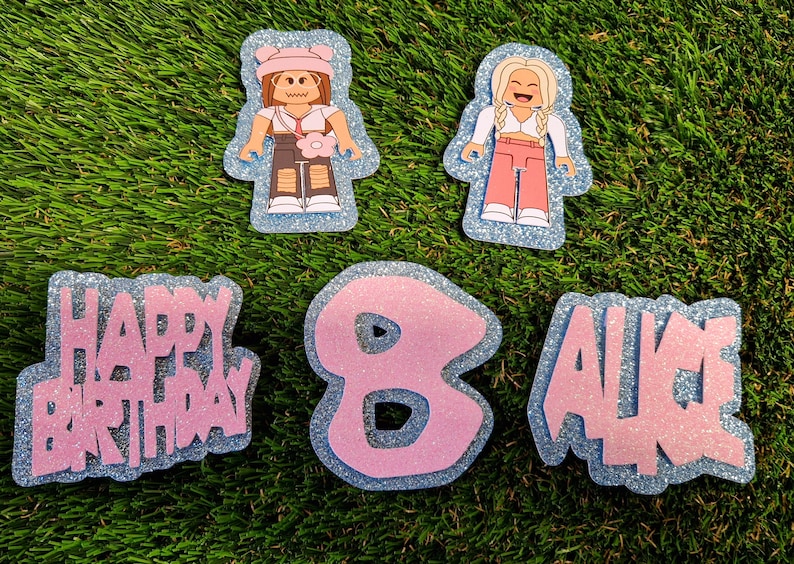 May include: A set of pink and blue glitter cake toppers featuring two cartoon figures, the words "Happy Birthday", the number "8", and the name "Alice".