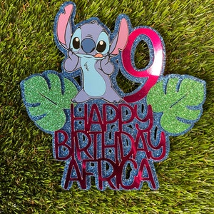 K&ouml;nnte beinhalten: Ein glitzernder blau-roter Tortenaufsatz zum Geburtstag mit der Figur Stitch aus Lilo & Stitch. Der Aufsatz enth&auml;lt die Zahl 9, gr&uuml;ne Bl&auml;tter und den Text "HAPPY BIRTHDAY AFRICA" in roter Glitzer-Schrift.