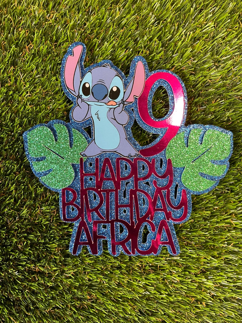 K&ouml;nnte beinhalten: Ein glitzernder blau-roter Tortenaufsatz zum Geburtstag mit der Figur Stitch aus Lilo & Stitch. Der Aufsatz enth&auml;lt die Zahl 9, gr&uuml;ne Bl&auml;tter und den Text "HAPPY BIRTHDAY AFRICA" in roter Glitzer-Schrift.