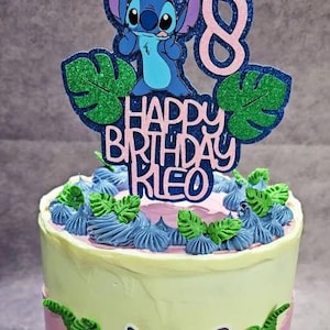K&ouml;nnte beinhalten: Ein Geburtstagskuchen mit einem Stitch-Themen-Topper. Der Topper zeigt eine blaue Stitch-Figur, gr&uuml;ne Glitzerbl&auml;tter und den Text "HAPPY BIRTHDAY KIE" in Rosa und Blau. Der Kuchen ist mit blauen und gr&uuml;nen Zuckerguss verziert.