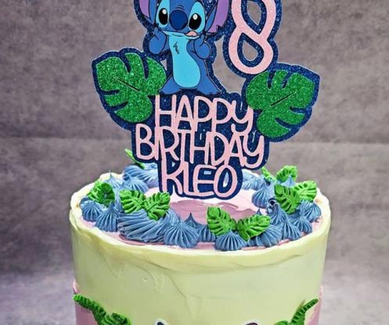 K&ouml;nnte beinhalten: Ein Geburtstagskuchen mit einem Stitch-Themen-Topper. Der Topper zeigt eine blaue Stitch-Figur, gr&uuml;ne Glitzerbl&auml;tter und den Text "HAPPY BIRTHDAY KIE" in Rosa und Blau. Der Kuchen ist mit blauen und gr&uuml;nen Zuckerguss verziert.